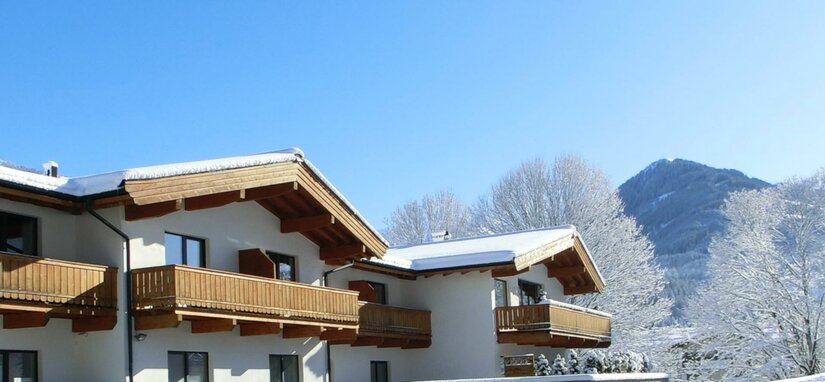 Winteransicht Kitz Mountain Living Alpines Gebäude mit Holzbalkonen und schneebedeckten Dächern vor blauem Himmel und Bergen.