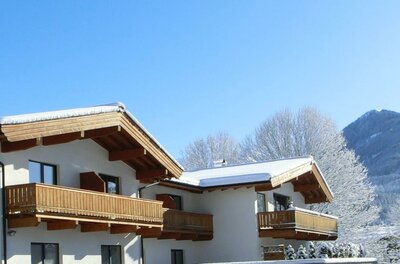 Alpines Gebäude mit Holzbalkonen und schneebedeckten Dächern vor blauem Himmel und Bergen.