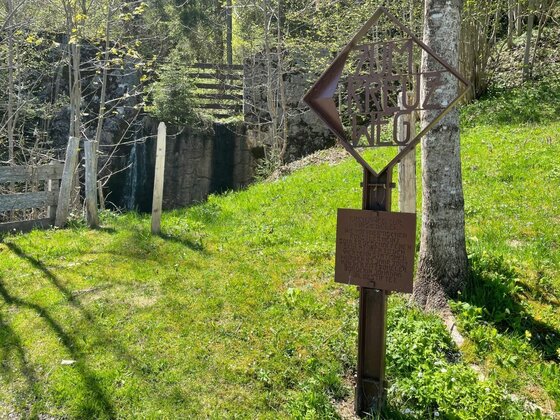 Beschilderung am Kreuzweg Ein rostfarbenes Metallschild mit der Aufschrift Kreuzweg auf einer grünen Wiese vor einer Steinmauer.