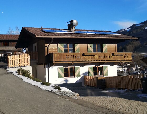 Außenansicht Haus Ingrid Zweistöckiges Alpenhaus mit Holzbalkon, grünen Fensterläden und Solaranlage auf dem Dach unter blauem Himmel.