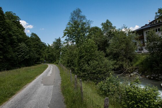 Wanderweg am Bachlauf Asphaltierter Wanderweg neben einem Bachlauf und Bäumen unter blauem Himmel.