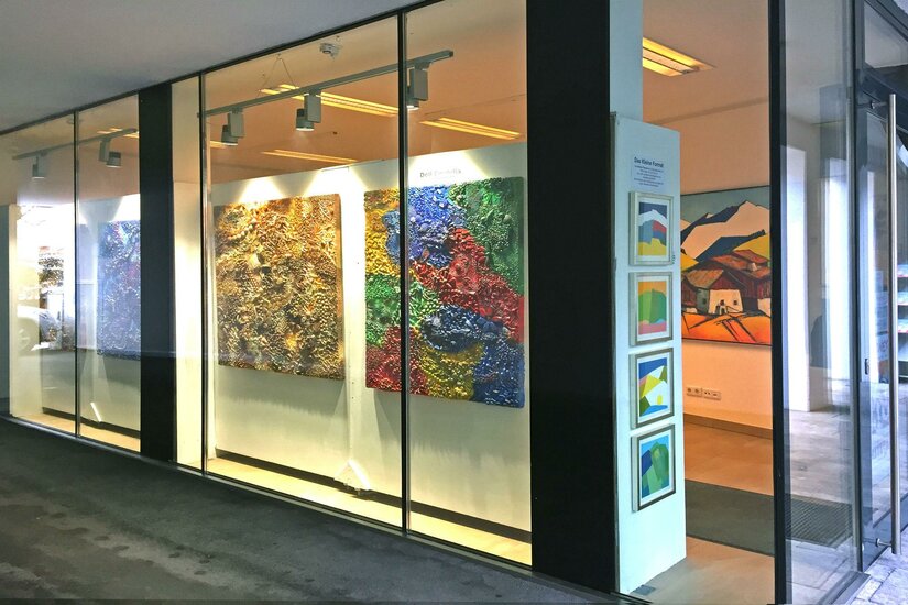 Galerie Kitz Art Eine Kunstgalerie mit großen, farbenfrohen Gemälden an der Wand. Die Fenster sind klar, und es gibt zusätzliche Kunstwerke in Rahmungen auf der rechten Seite.
