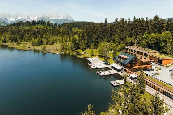 Steghaus am See Luftaufnahme eines Holzgebäudes mit Steganlagen an einem See vor bewaldeten Hügeln und Gebirge.
