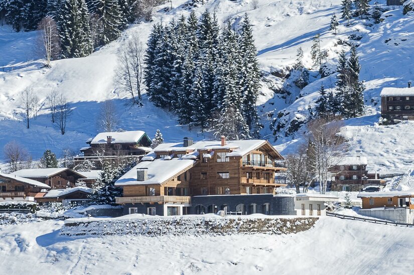 KitzChalet Jochberg im Winter Mehrstöckiges Holzchalet mit schneebedecktem Dach in einer alpinen Winterlandschaft vor bewaldetem Hang.