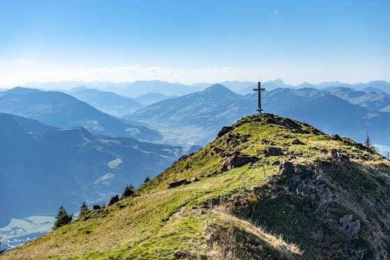 Ein hölzernes Gipfelkreuz auf einem grasbewachsenen Bergrücken vor einem Panorama aus blauen Gebirgsketten.
