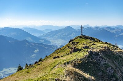 Gipfelkreuz am Stuckkogel Ein hölzernes Gipfelkreuz auf einem grasbewachsenen Bergrücken vor einem Panorama aus blauen Gebirgsketten.