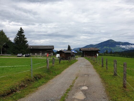 Jagahäusel bei Westendorf Schotterweg führt zu mehreren Holzhütten auf einer grünen Wiese vor bewaldeten Bergen unter bewölktem Himmel.