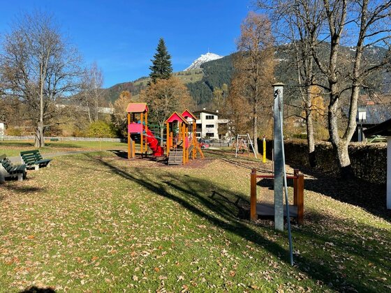 Spielplatz Pfarrau Kinderspielplatz mit Klettergerüst, Rutsche und Schaukeln vor bewaldeten Bergen unter blauem Himmel.