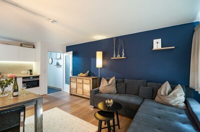 Modernes Wohnzimmer mit blauem Ecksofa, Holztisch und Küchenzeile im Hintergrund.