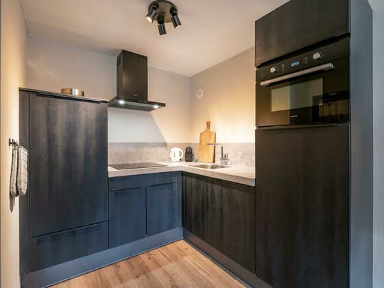 Küche im Appartement Hafkamp Moderne dunkle Küchenzeile über Eck mit Backofen, Kochfeld und Spüle in einem Appartement.