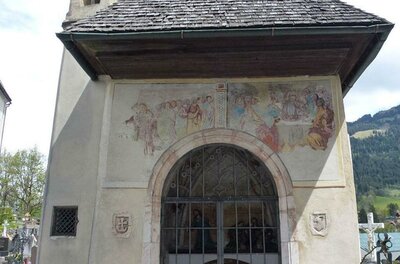 Eine kleine Kapelle mit einem schrägen Dach und einem Gittertor. An der Fassade sind alte Fresken sichtbar.