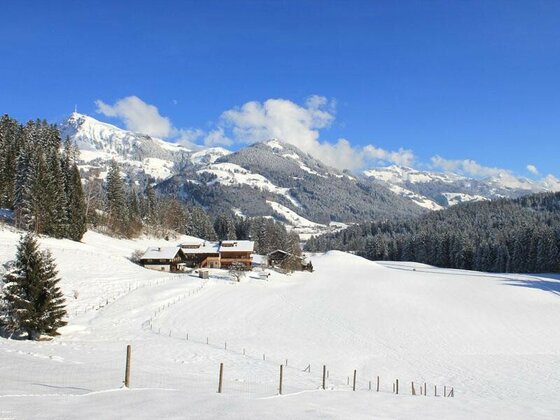 Verschneite Berglandschaft mit Holzhäusern, Nadelwald und schneebedeckten Gipfeln unter blauem Himmel.