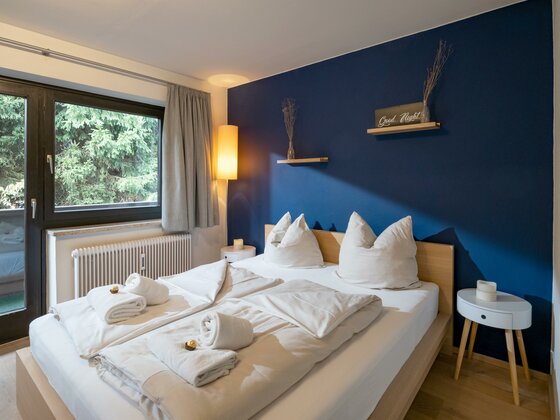 Modernes Doppelbett vor einer dunkelblauen Wand mit Holzregalen und Fenster zum Wald.