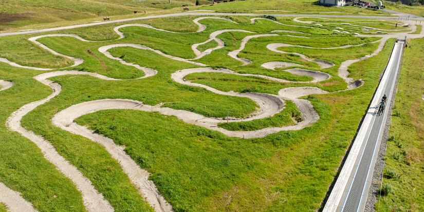Eine kurvenreiche Strecke aus Schotter und Gras im MTB Family Area bei den 360 Flow Trails in Kitzbühel führt über eine grüne Wiese, umgeben von sanften Hügeln und Bäumen.