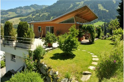Modernes Holzhaus mit Pultdach und Garten vor bewaldeten Bergen.