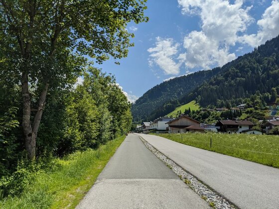 Asphaltierter Rad- und Gehweg neben einer Straße vor bewaldeten Bergen und Häusern.