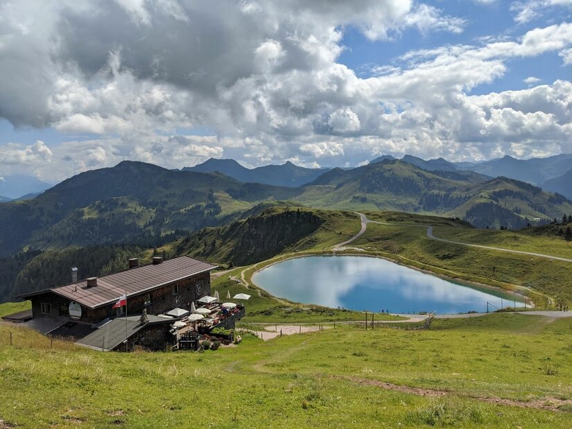 Berghütte mit Terrasse neben einem runden Speichersee in einer alpinen Graslandschaft vor Gebirgskette.