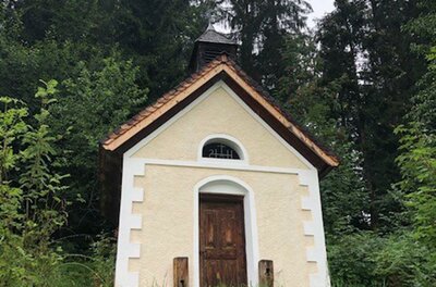 Eine kleine Kapelle inmitten von dichten Bäumen. Das Gebäude hat eine hellbeige Fassade und ein steiles Dach.