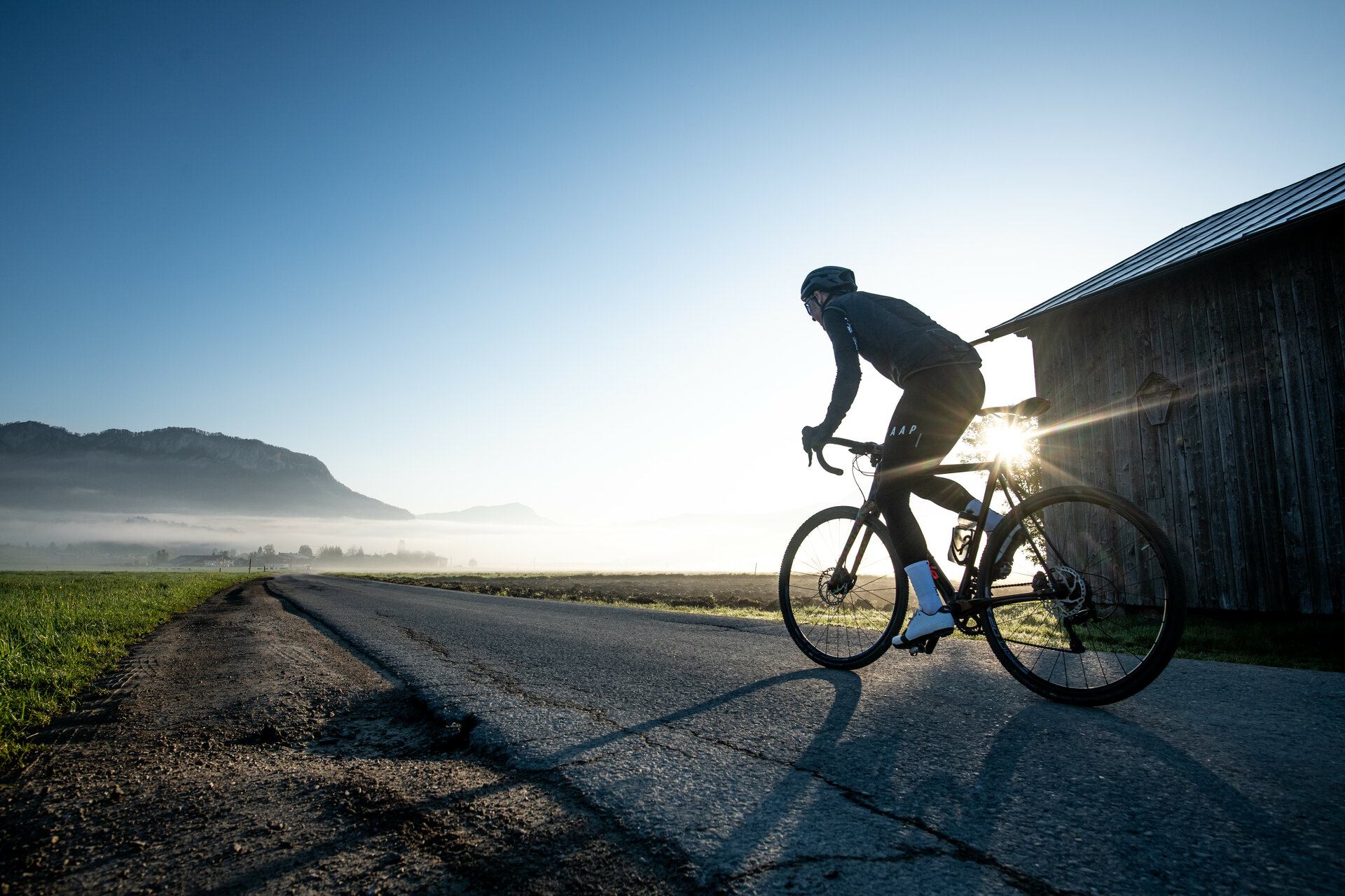 Ein Radfahrer fährt auf einer ruhigen Straße, umgeben von Nebel und Bergen, während die Sonne aufgeht.
