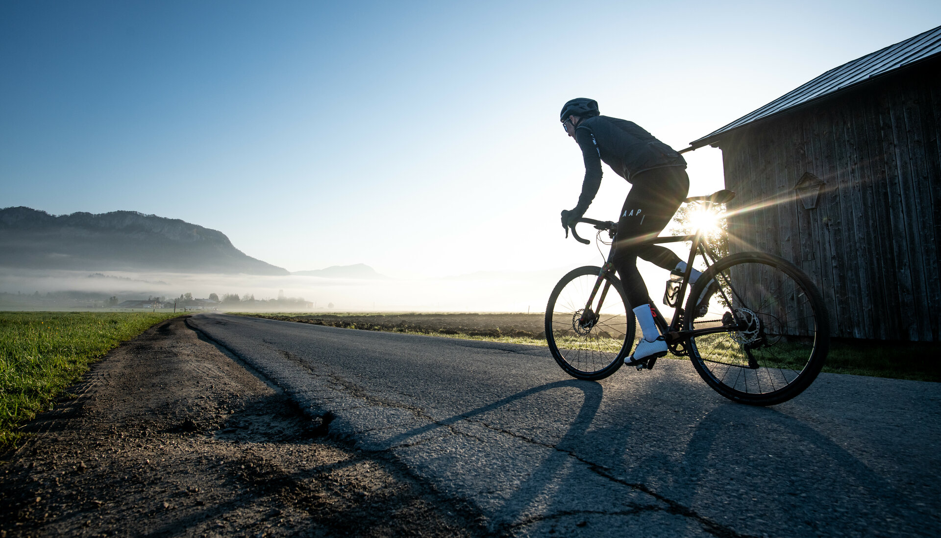 Ein Radfahrer fährt auf einer ruhigen Straße, umgeben von Nebel und Bergen, während die Sonne aufgeht.