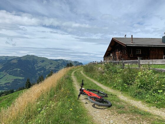 Ein rotes Mountainbike liegt auf einem Schotterweg neben einer Holzhütte vor Bergpanorama.