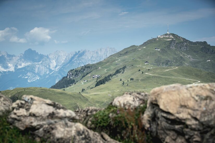 Blick auf das Kitzbüheler Horn mit Sendemast, Wanderwegen und Almhütten vor felsiger Gebirgskulisse.