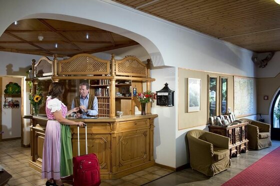 Rezeption Hotel Wiesenegg Frau im Dirndl mit Koffer an einer hölzernen Hotelrezeption im Gespräch mit einem Mitarbeiter.