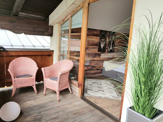 Zwei rosa Korbsessel auf einem Holzbalkon neben einer Glasfront zum Innenraum.