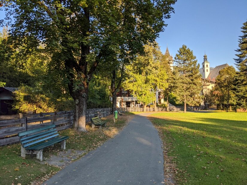 Asphaltierter Fußweg in einer Parkanlage mit Holzbänken, Bäumen und einer Kirche im Hintergrund.