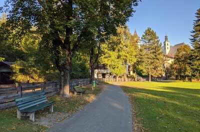 Asphaltierter Fußweg in einer Parkanlage mit Holzbänken, Bäumen und einer Kirche im Hintergrund.