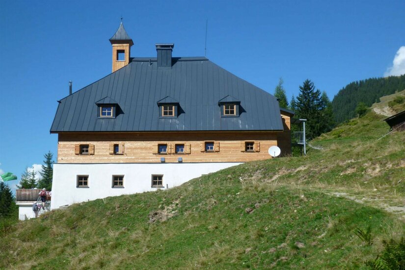Kelchalm im Sommer Dreistöckiges alpines Gebäude mit dunklem Metalldach und kleinem Glockenturm auf einem Grashang.