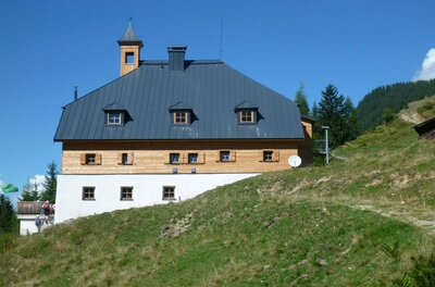 Dreistöckiges alpines Gebäude mit dunklem Metalldach und kleinem Glockenturm auf einem Grashang.