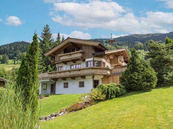 Mehrstöckiges Alpenhaus aus Holz und weißem Putz mit Balkonen auf einer grünen Wiese vor bewaldeten Bergen.