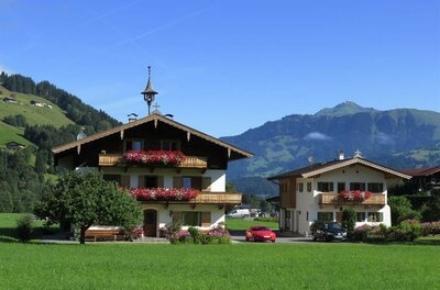 Zwei traditionelle Alpenhäuser mit Blumenschmuck vor einer Bergkulisse unter blauem Himmel.