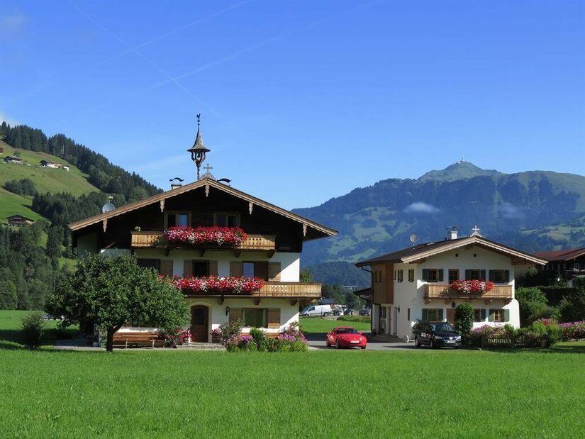 Zwei traditionelle Alpenhäuser mit Blumenschmuck vor einer Bergkulisse unter blauem Himmel.