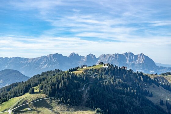 Blick auf bewaldete Bergrücken mit Gebäuden auf dem Gipfel vor einer schroffen Felskette unter blauem Himmel.