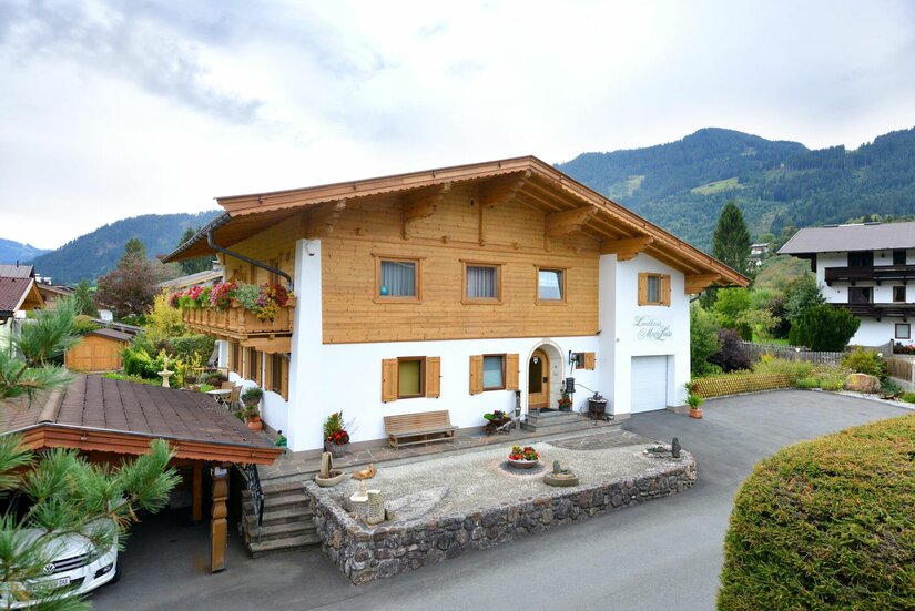 Zweistöckiges Landhaus mit Holzfassade im Obergeschoss und Bergen im Hintergrund.