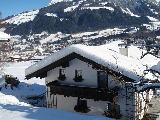 Verschneites Haus mit Holzbalkon vor einer alpinen Bergkulisse und einer Ortschaft im Tal.