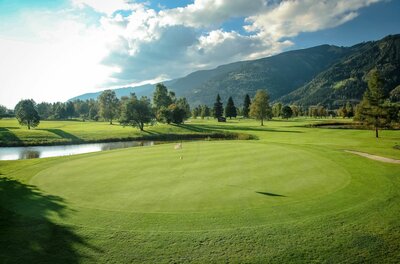 Golfclub Zell am See Gepflegtes Green auf einem Golfplatz mit Wasserhindernis vor bewaldeten Bergen unter bewölktem Himmel.
