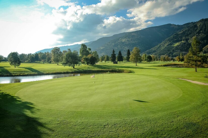 Golfclub Zell am See Gepflegtes Green auf einem Golfplatz mit Wasserhindernis vor bewaldeten Bergen unter bewölktem Himmel.