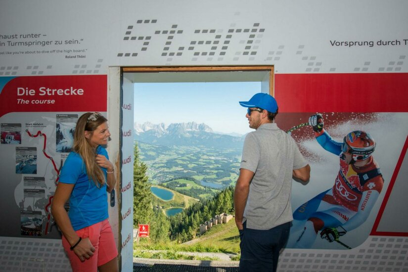 Starthütte Ein Blick durch eine Tür auf eine atemberaubende Landschaft mit Bergen und Tälern. Zwei Personen stehen davor und unterhalten sich.