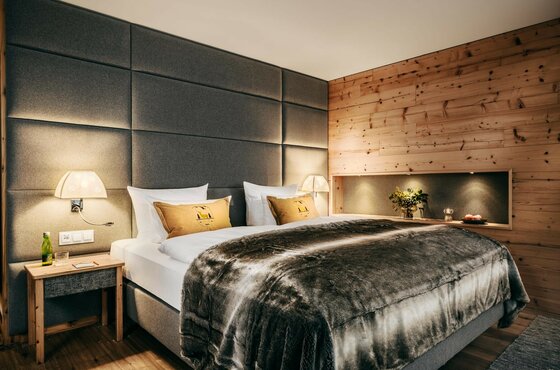 Grand Tirolia Zimmer Doppelbett mit grauer Polsterwand, Pelzdecke und Holzverkleidung in einem Hotelzimmer.