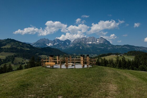 Eingezäunter Aussichtspunkt mit Holzbank auf einem Grashügel vor einem Gebirgspanorama unter blauem Himmel.