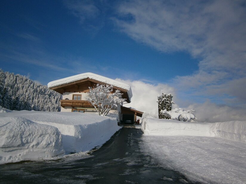 Haus Jöchl in Reith Alpines Wohnhaus mit schneebedecktem Dach und geräumter Zufahrt vor bewaldetem Hang unter blauem Himmel.