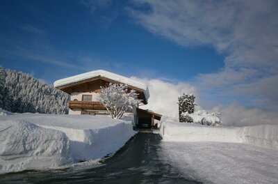 Haus Jöchl in Reith Alpines Wohnhaus mit schneebedecktem Dach und geräumter Zufahrt vor bewaldetem Hang unter blauem Himmel.