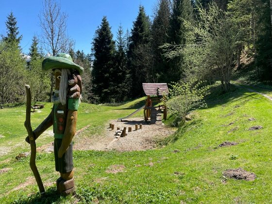 Spielplatz Einsiedelei Holzfigur eines Wanderers auf einer Wiese vor einem Waldspielplatz mit Rutsche.