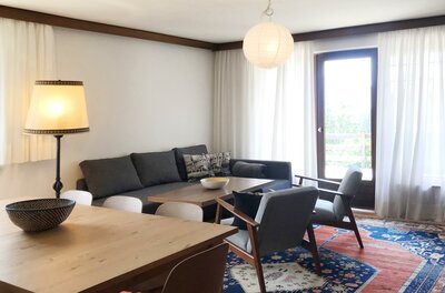 Wohnzimmer Hahnenkamm Wohnzimmer mit grauem Sofa, zwei Sesseln, Holztisch und buntem Teppich vor einer Balkontür.