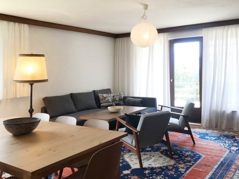 Wohnzimmer Hahnenkamm Wohnzimmer mit grauem Sofa, zwei Sesseln, Holztisch und buntem Teppich vor einer Balkontür.
