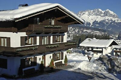 Alpines Gästehaus mit Holzbalkonen und schneebedecktem Dach vor verschneitem Bergmassiv.