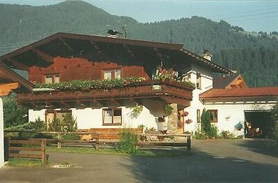Alpines Wohnhaus mit Holzbalkon und Blumenschmuck vor bewaldetem Berg.